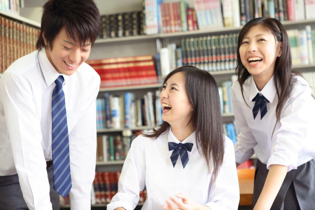 図書館の机を囲んで笑い合う高校生たち。男子学生と女子学生2人が明るい表情で会話している様子。