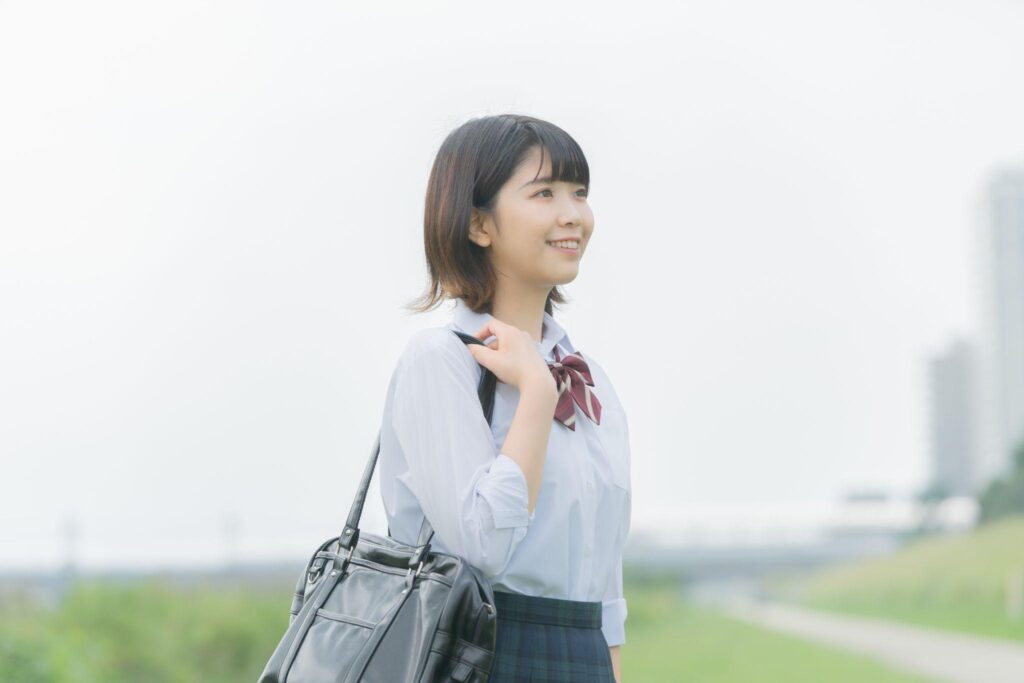 屋外で通学バッグを肩にかけ、前を向いて微笑む女子高校生。